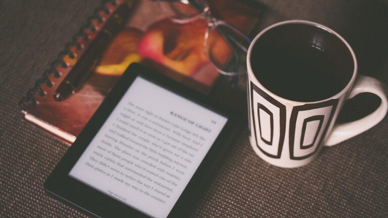 An e-reader placed next to a mug, reading glasses and notebook and pen
